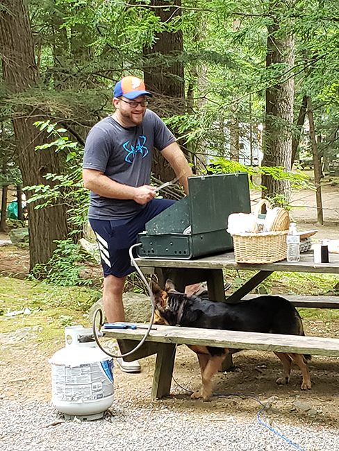 Man cooking breakfast on a Coleman camping stove with a dog waiting for food droppings